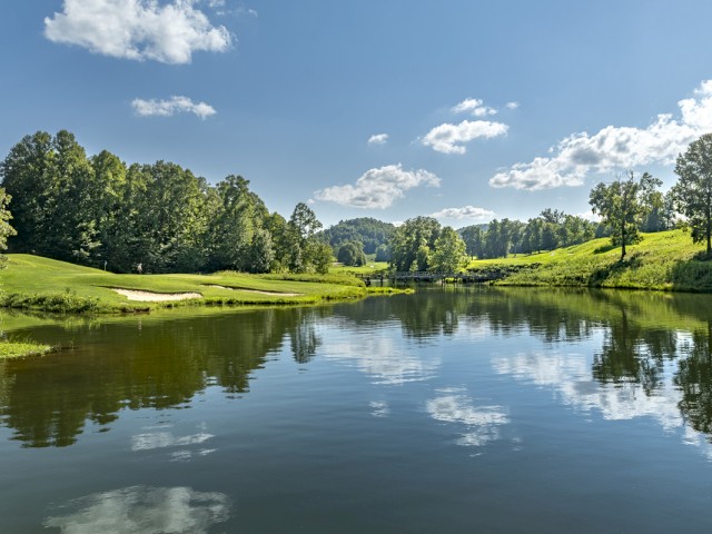 A peaceful lakeside landscape with clear blue skies, fluffy white clouds, and a lush green forest reflecting on the water's surface.