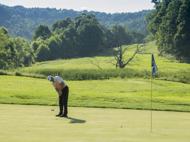 A person is swinging a golf club on a green, with the golf flag and hole nearby, surrounded by a lush, hilly landscape and trees.