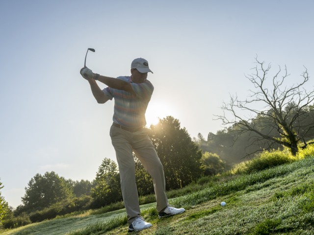 A person is preparing to swing a golf club on a grassy course, with the sun rising behind them and trees in the background, ending the sentence.