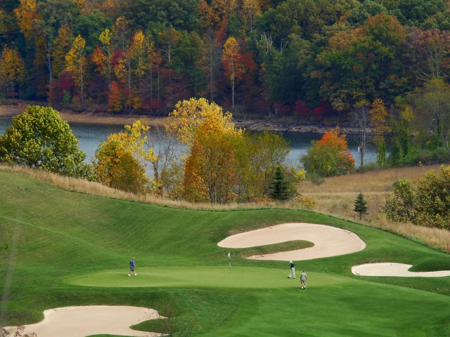 A scenic golf course in autumn with players on the green, surrounded by trees with colorful foliage, and a lake in the background.