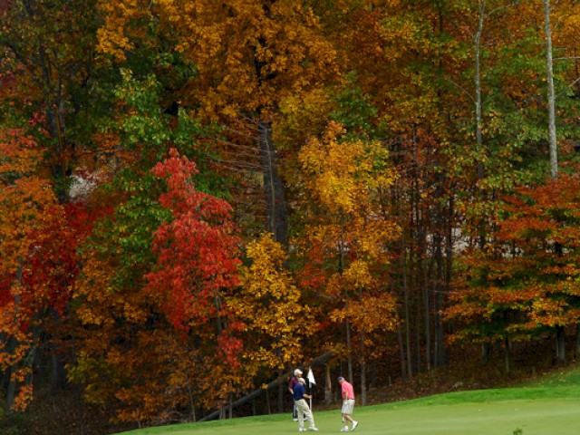 Two people are playing golf on a green course surrounded by autumn foliage with trees displaying vibrant red, yellow, and orange leaves.