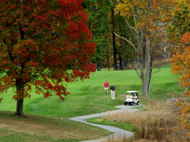 A scenic autumn golf course with vibrant red and orange trees; two people and a golf cart are visible in the background near a winding path.