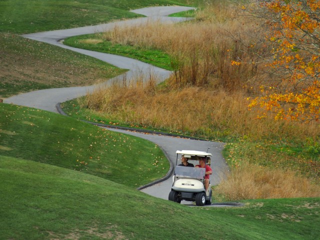 A golf cart with two people is driving on a winding path through a golf course with lush green grass and some autumn trees in the background.