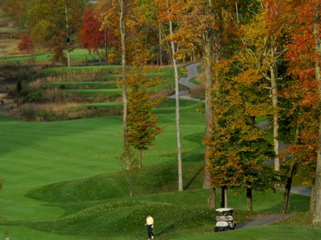 The image shows a golf course during autumn, with a golfer near the hole and a golf cart nearby, surrounded by colorful trees.