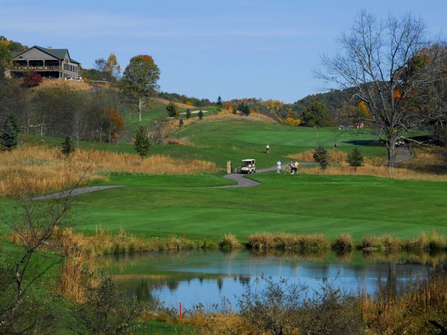 Golf course with lush green fairways, a pond in the foreground, a golf cart, and people playing on a sunny day, with a house on a hill.