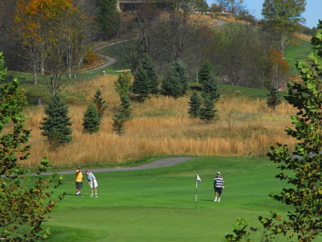 A picturesque golf course scene with three players on the green, surrounded by trees and a scenic hilltop house in the background, ending the sentence.