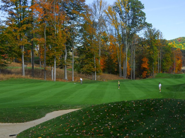 A golf course with three players on the green, autumn trees in the background, and a sand bunker in the foreground, ending the sentence.
