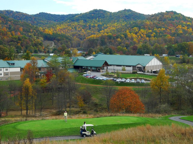 A scenic view of a golf course with green hills in the background and buildings surrounded by autumn trees.
