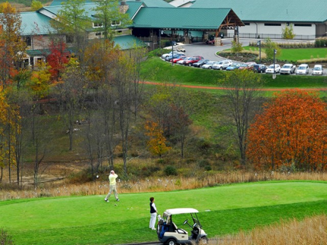 A scenic autumn landscape featuring a golf course with players and a golf cart, and a large building in the background surrounded by trees and hills.
