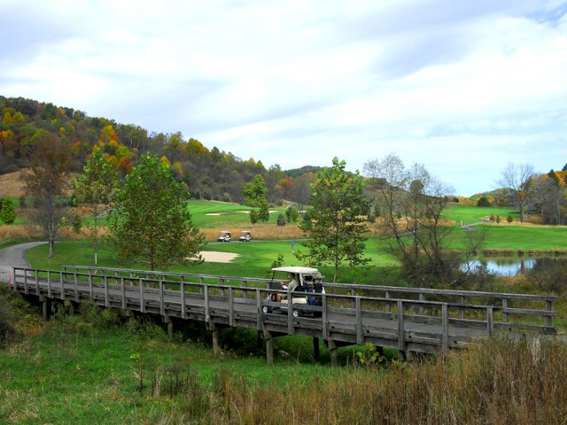A wooden bridge over grassy terrain with a golf cart, surrounded by green golf course and autumn-colored trees, under a partly cloudy sky.
