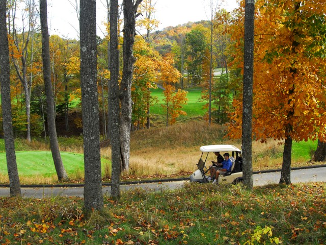 A golf cart is seen driving along a path through a scenic, autumnal landscape with colorful trees and a grassy area.