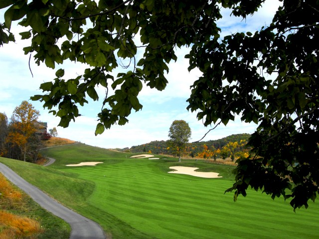 A scenic view of a green golf course with sand bunkers, a winding pathway, trees in the background, and foliage framing the image.