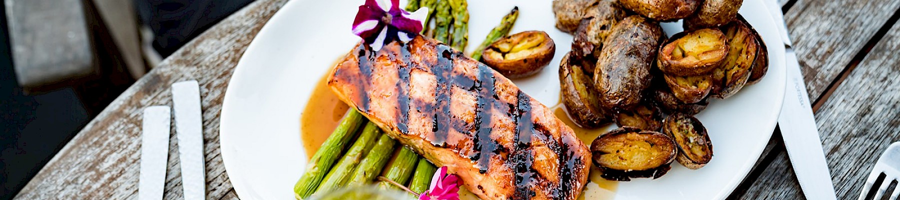 A beautifully plated meal with grilled salmon, roasted potatoes, asparagus, and a glass of white wine on a rustic wooden table.