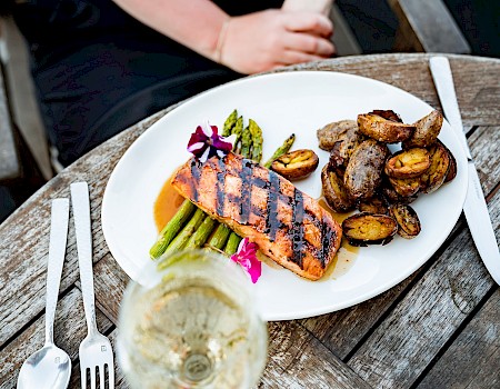 A beautifully plated meal with grilled salmon, roasted potatoes, asparagus, and a glass of white wine on a rustic wooden table.
