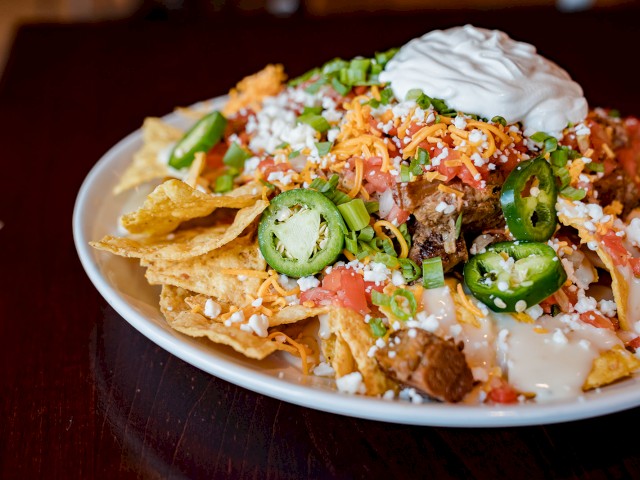 A plate of loaded nachos topped with jalapeños, cheese, tomatoes, green onions, sour cream, and other toppings on a dark wooden table.