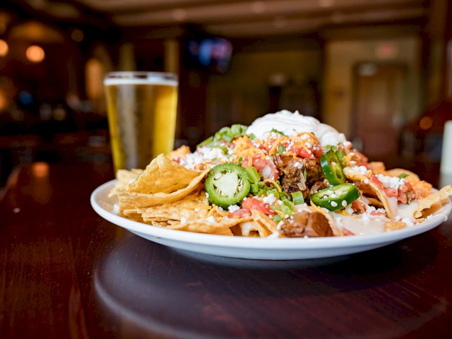 A plate of nachos topped with jalapeños, melted cheese, sour cream, and other toppings sits next to a glass of beer on a wooden table.