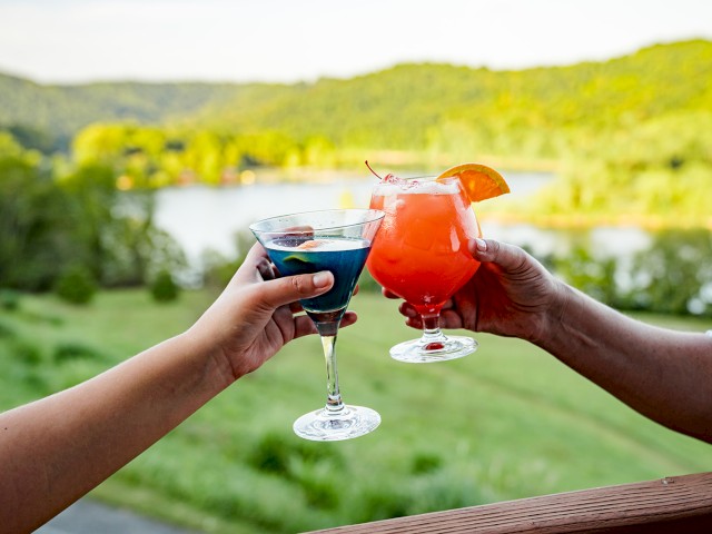 Two people clink glasses against a scenic backdrop of greenery and a body of water, with one holding a blue drink and the other a red drink.