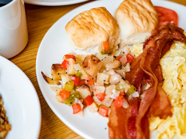 A breakfast plate with scrambled eggs, bacon, roasted potatoes, and biscuits. A cup of coffee and another dish are partially visible.