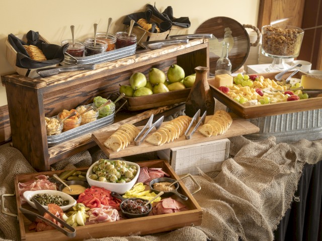 The image shows a rustic buffet with assorted fruits, cheeses, bread, olives, nuts, crackers, and condiments on a burlap-covered table.