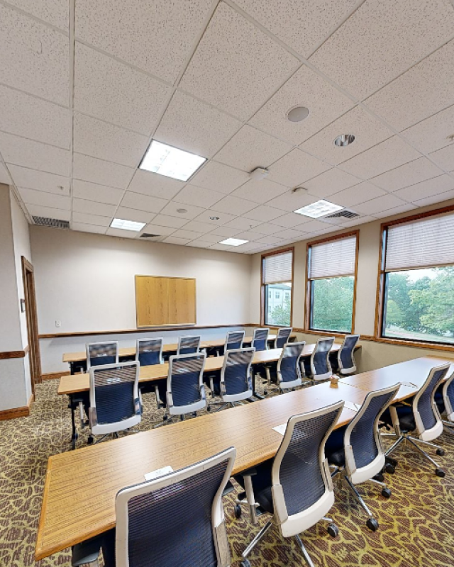This image shows an empty conference or meeting room with desks, chairs, windows, and a wall with a whiteboard.