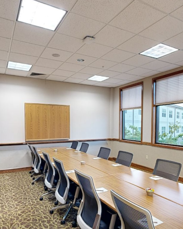 Empty conference room with a long table, chairs, papers, and small bowls, surrounded by windows showing trees and buildings outside.