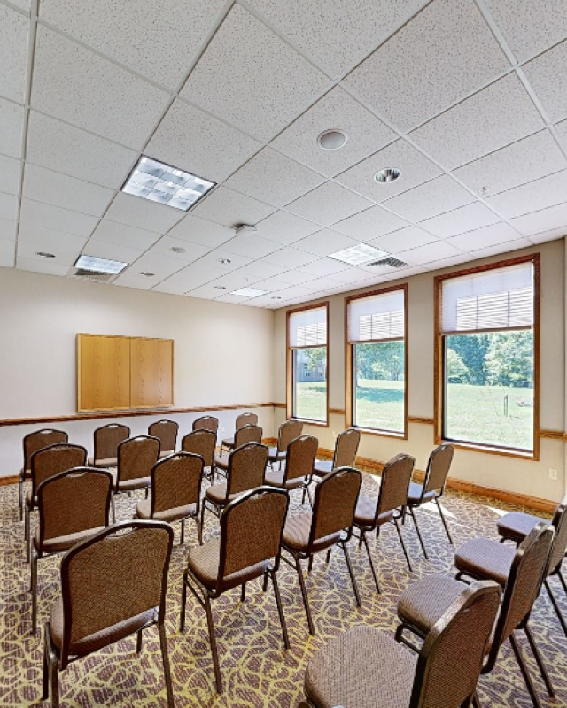 An empty conference or meeting room with chairs arranged in rows, large windows, and a cabinet near a decorated ceiling.