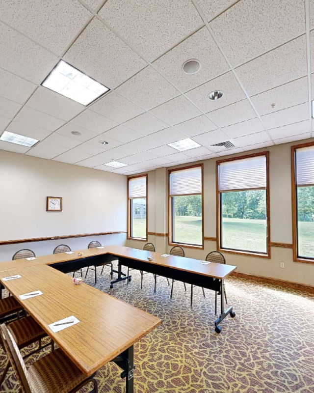 A bright conference room with a U-shaped table setup, large windows showing greenery, and ceiling lights in a professional space.