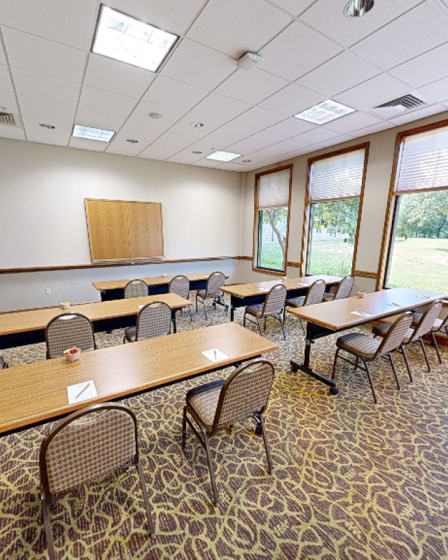 An empty conference or meeting room with tables, chairs, large windows, and a carpeted floor.