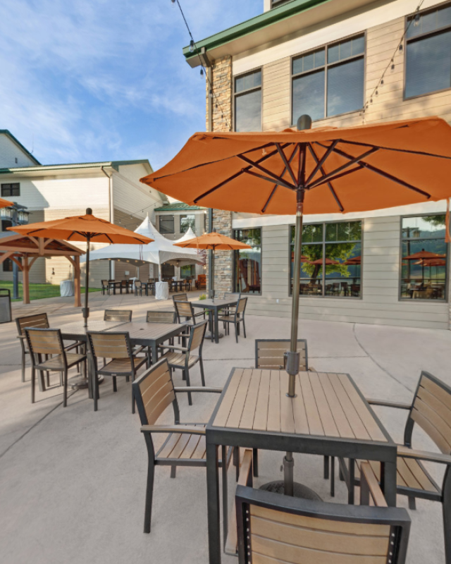 Empty outdoor patio with tables, chairs, orange umbrellas, and a brick and wood building in the background.