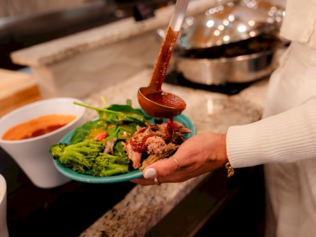 A person is pouring sauce over a plate of cooked vegetables and meat in a kitchen setting.