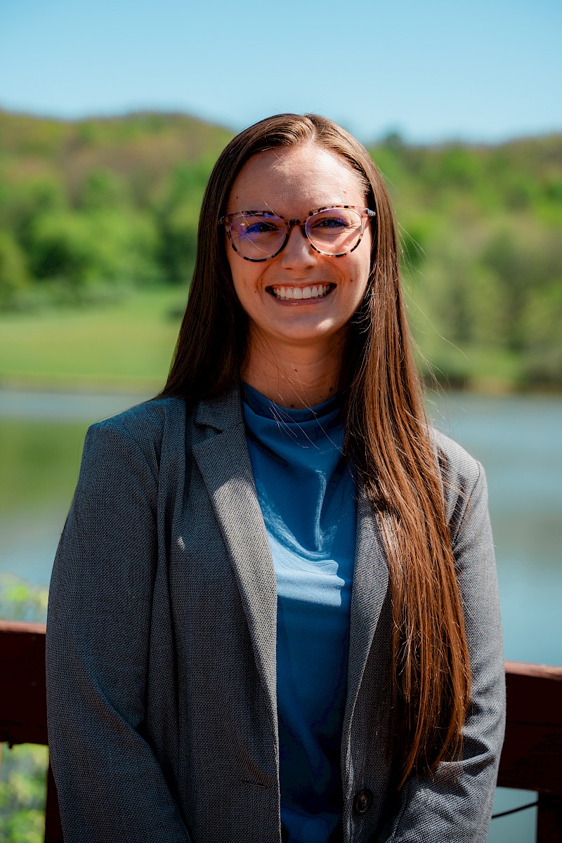 A woman with long brown hair and glasses, smiling outdoors near a lake with green trees in the background.