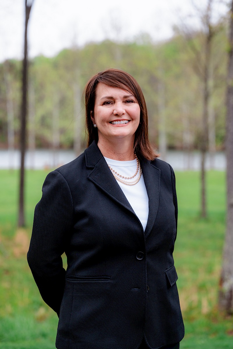 A woman in a black blazer and white top is smiling outdoors near a lake with trees in the background.