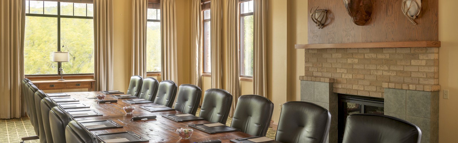 A conference room with a long wooden table, black chairs, a fireplace, deer heads mounted above, and large windows with beige curtains.