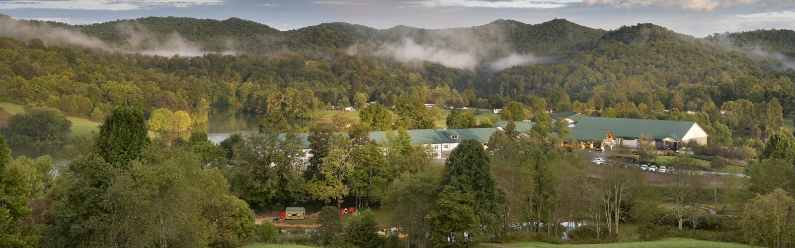 A scenic view of green hills with scattered trees, a few buildings, and a meandering road under a partly cloudy blue sky.