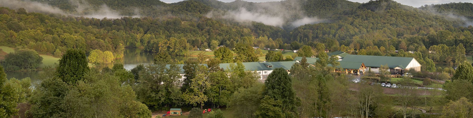 A scenic view of green hills with scattered trees, a few buildings, and a meandering road under a partly cloudy blue sky.