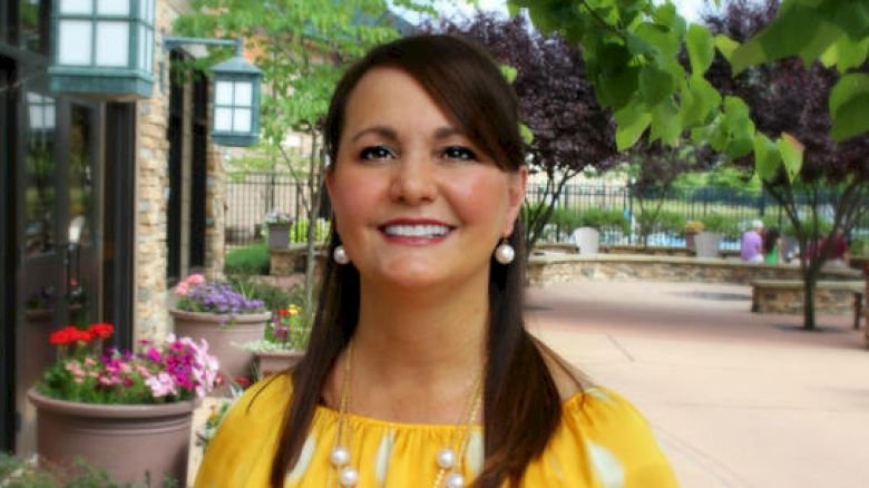 A woman in a yellow top and pearl necklace is smiling outdoors near flowers and greenery, with a building in the background.