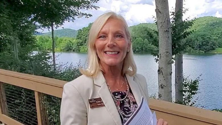 A smiling woman stands on a wooden deck holding papers, with a lake and trees in the background under a partly cloudy sky.