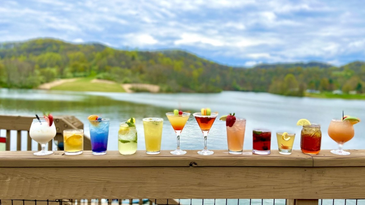 A variety of colorful drinks are lined up on a wooden railing with a scenic lake and mountains in the background under a partly cloudy sky.