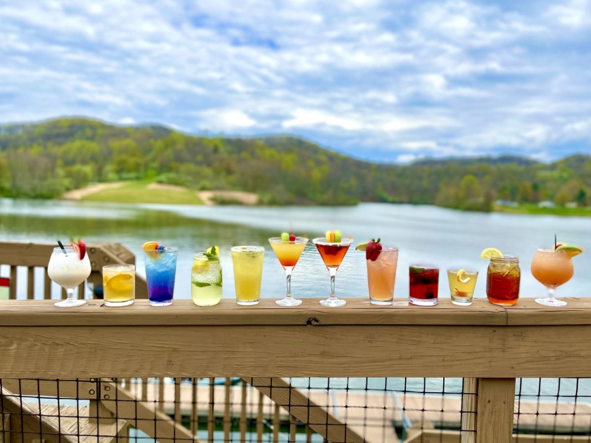 A variety of colorful drinks are lined up on a wooden railing with a scenic lake and mountains in the background under a partly cloudy sky.