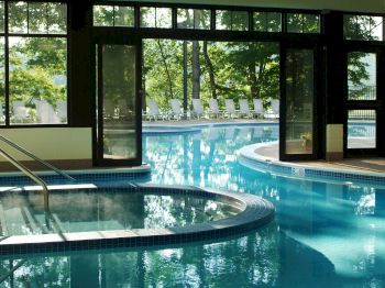 A serene indoor-outdoor pool area with clear blue water, surrounded by glass walls and doors, which open to a view of trees and pool chairs outside.