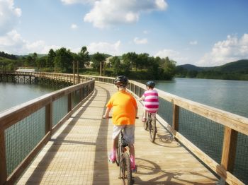 Two people riding bicycles on a wooden path over a lake, surrounded by trees and hills under a partly cloudy sky.