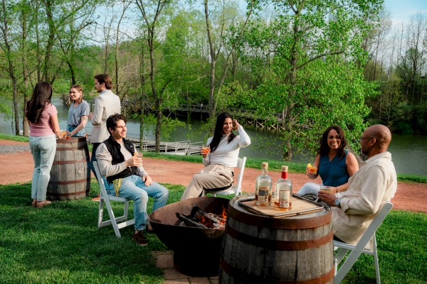 A group of friends enjoys a barbecue near a river on a sunny day, relaxing, chatting, and sipping drinks by the fire.