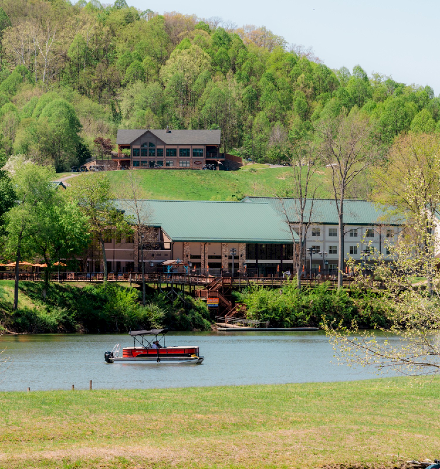 A boat floats on a calm river with a green hillside and buildings, surrounded by trees in a peaceful countryside scene.