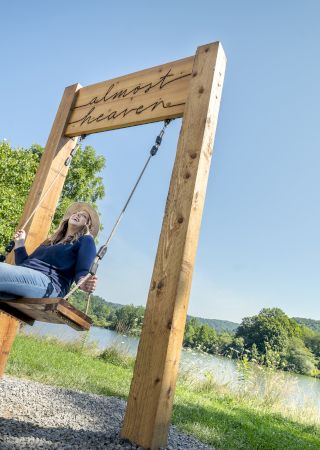 A person is joyfully sitting on a large swing in a picturesque lakeside park with greenery and clear skies. The swing has 