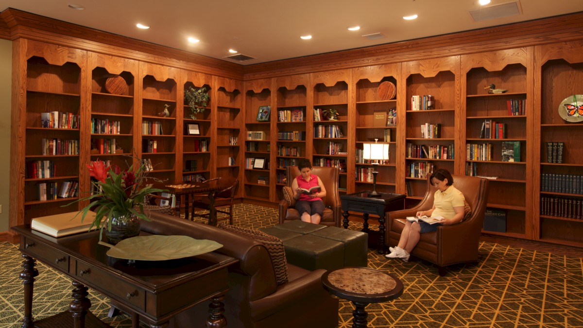 A cozy library room with wooden bookshelves, tables, and chairs. Two people are seated and reading books in comfortable armchairs.