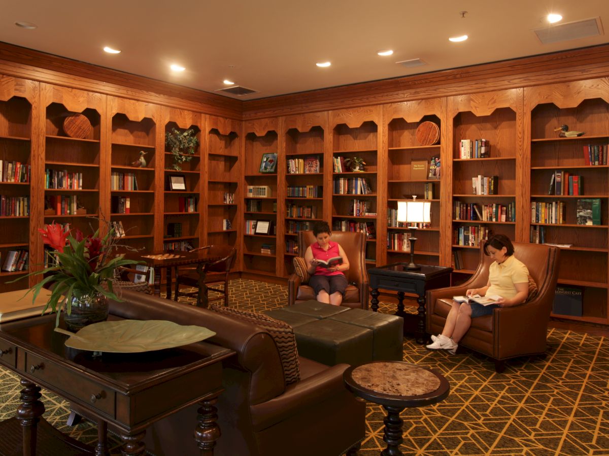 A cozy library room with wooden bookshelves, tables, and chairs. Two people are seated and reading books in comfortable armchairs.
