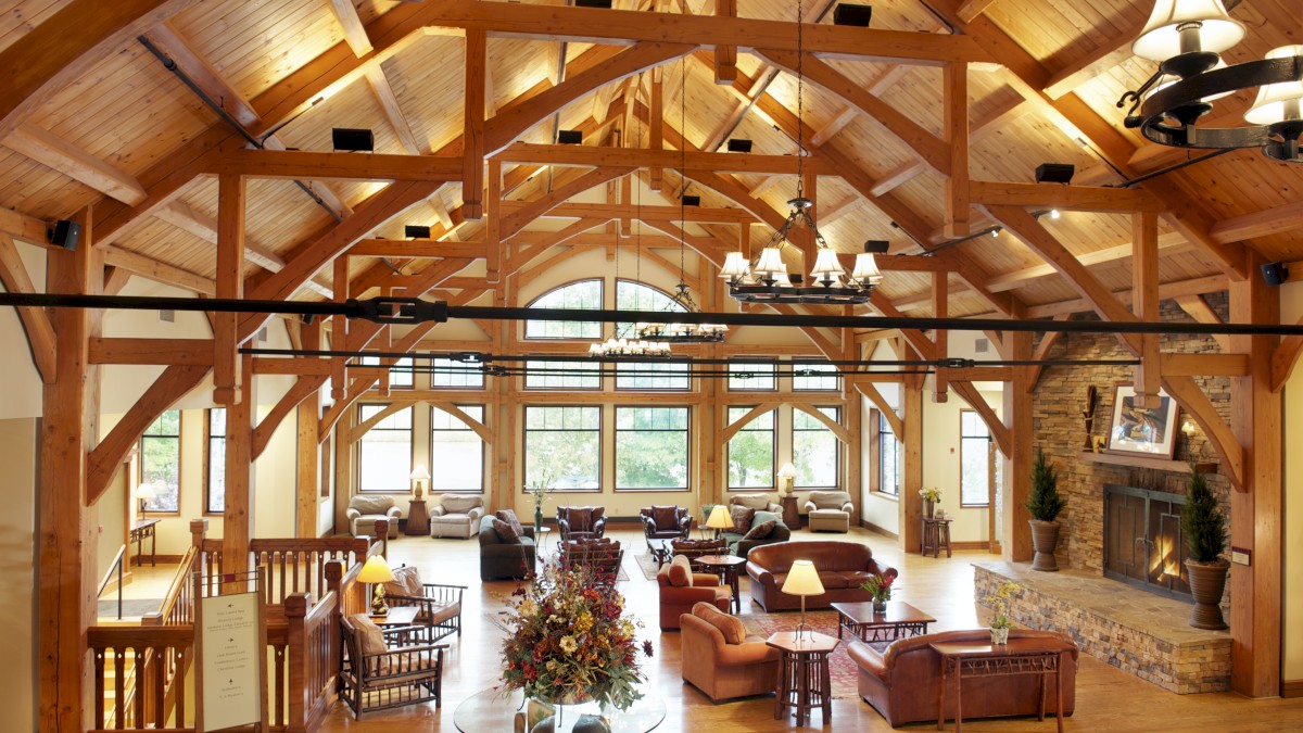 The image shows a large, rustic lodge interior with wooden beams, cozy seating areas, a stone fireplace, chandeliers, and natural light from large windows.