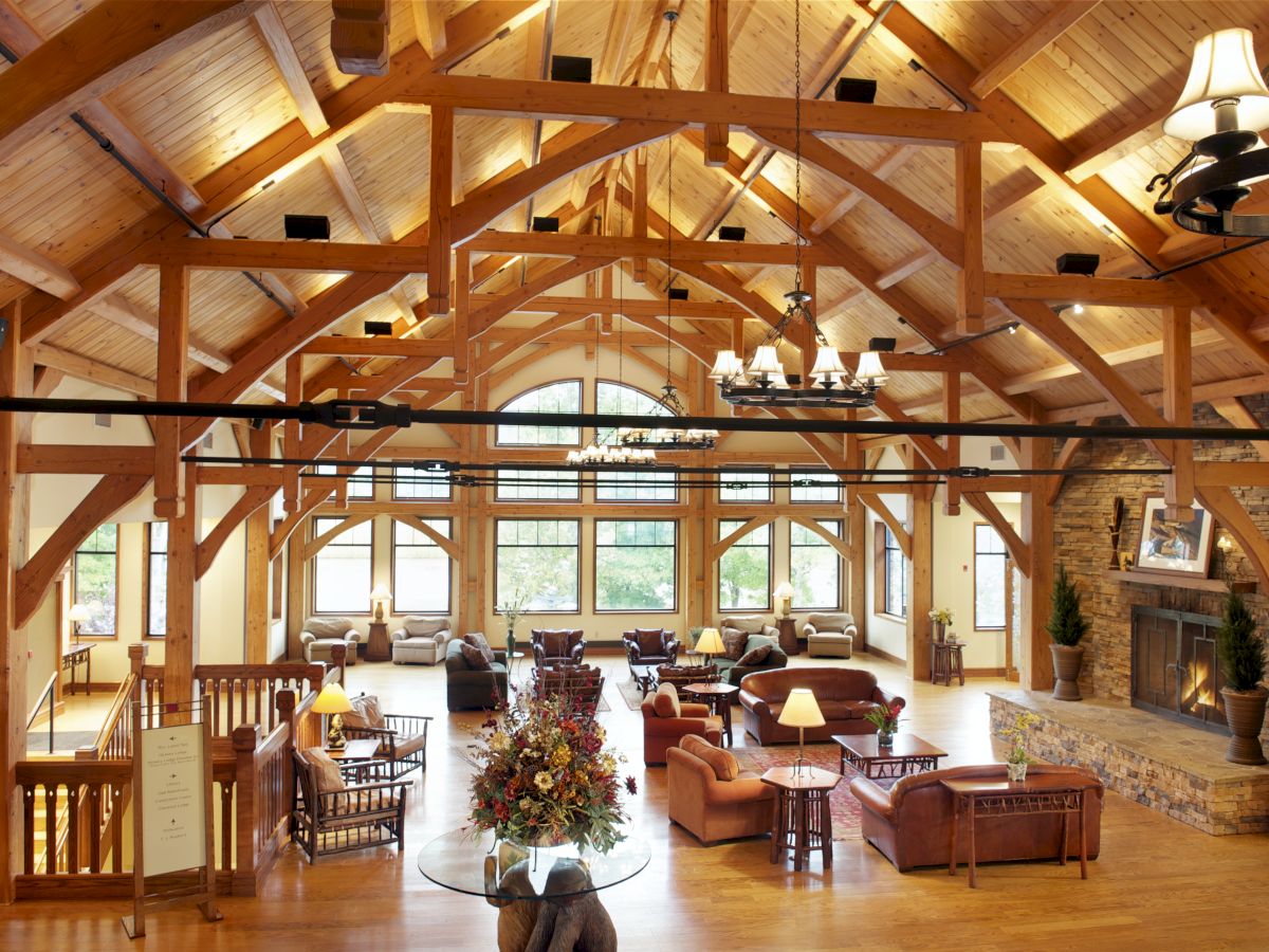 The image shows a large, rustic lodge interior with wooden beams, cozy seating areas, a stone fireplace, chandeliers, and natural light from large windows.