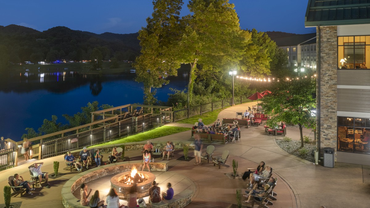 People are sitting around a fire pit near a lake at dusk, surrounded by trees and lights. The area is part of a larger complex and looks relaxing.