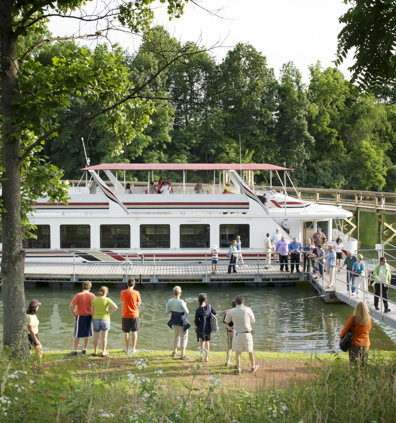 A group of people are boarding a white boat via a dock surrounded by greenery and trees. The scene is lively and looks peaceful.